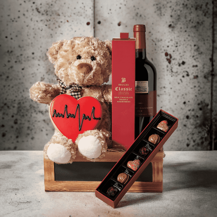 "I Love You Too" Wine & Chocolate Basket includes a Tan Plush Snuggle Bear, Bottle of Wine, Heart Sugar Cookie With Royal Icing, Truffles, and a Small Slate Front Wooden Box.
